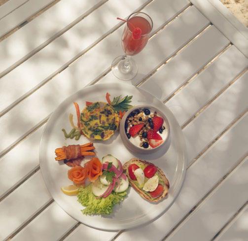 A top-down view of a white plate on a white slatted table. The plate features a vegetable frittata, a bowl of yogurt with berries and granola, smoked salmon rosettes, and avocado toast. A tall glass of reddish mimosa with a cherry sits next to the plate in the sunlight.