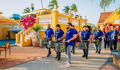 A group of male drummers in matching blue polo shirts and sunglasses marching along a paved walkway. They are playing colorful, decorated drums. In the background, a person in a bright pink carnival costume stands on a yellow balcony under a clear sky with palm trees.