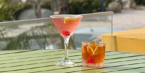 A close-up of two cocktails sitting on a green slatted table outdoors. One is a pink martini-style drink with a lemon twist and cherry, and the other is an amber-colored short drink with a heart-shaped orange peel garnish. The background is softly blurred with hints of tropical greenery.