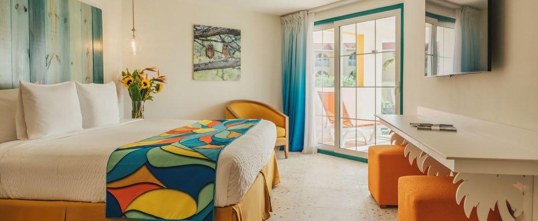 A brightly lit hotel room featuring a large bed with a colorful, abstract-patterned runner in blue, orange, and green. The room includes a teal wooden headboard, a white desk with orange stools, and sliding glass doors that open to a sunlit patio. A painting of two owls hangs on the wall, and a bouquet of sunflowers sits on a side table.